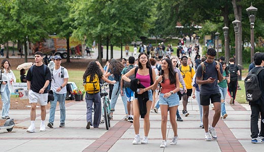 Georgia Tech students milling about the large sidewalk bordering Tech Green, the central green space of campus between the student center and learning commons. The background is populated by many trees lining the walkway.